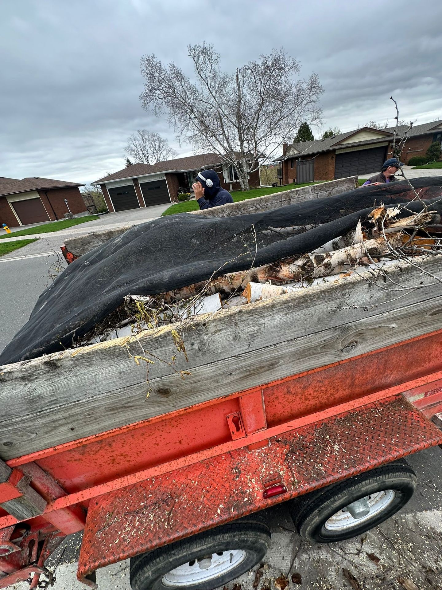 Red trailer filled with branches and debris covered by a tarp, parked on a residential street.